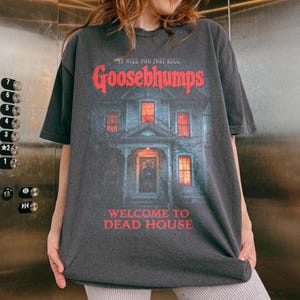 May include: Dark grey Goosebumps t-shirt with a haunted house graphic and red text: "IT WILL YOU JUST KILL Goosebumps WELCOME TO DEAD HOUSE". The tee has a vintage, worn appearance.