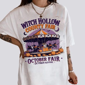 May include: White t-shirt featuring a Halloween-themed design. The graphic includes a carousel with witch figures, pumpkins, and the text "Witch Hollow County Fair" and "October Fair".