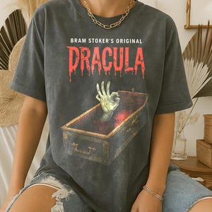 May include: Dark gray t-shirt featuring the text "Bram Stoker's Original Dracula" in red, dripping font. A green hand emerges from a coffin. The shirt has a vintage, worn look.