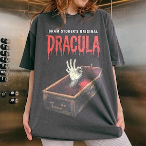 May include: Dark gray t-shirt featuring the text "BRAM STOKER'S ORIGINAL DRACULA" in red dripping font. The graphic shows a skeletal hand emerging from a coffin. The shirt has short sleeves and a crew neck.