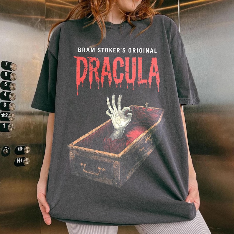 May include: Dark gray t-shirt featuring the text "BRAM STOKER'S ORIGINAL DRACULA" in red dripping font. The graphic shows a skeletal hand emerging from a coffin. The shirt has short sleeves and a crew neck.