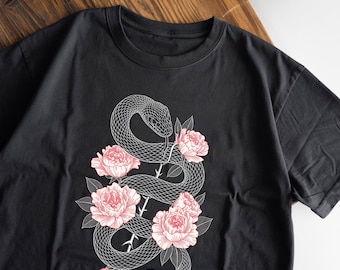May include: Black t-shirt featuring a white illustration of a snake intertwined with pink roses. The snake is detailed with scales, and the roses have multiple petals. The design is centered on the shirt, creating a striking contrast.