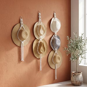 May include: Three white macrame wall hangers made of knotted cord, designed to hold hats. One hanger holds a single straw hat, while the other two hangers are vertical displays, each holding two hats of varying shades of tan, beige, and white.