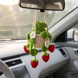 May include: A crocheted hanging plant ornament featuring a beige basket, green leaves, white flowers, and three red strawberries. The item is suspended by two beige yarn cords and is designed to hang from a vehicle rearview mirror.