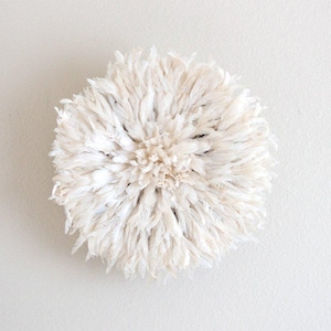 May include: A round, off-white wall hanging made of feathers. The feathers are arranged in a radial pattern, creating a textured, layered effect. The central area is denser, with a lighter hue.