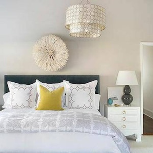May include: A bedroom scene featuring a bed with white bedding and decorative pillows. A yellow pillow sits in the center. A white nightstand with a lamp and a decorative chandelier are also visible. A decorative wall hanging is above the bed.