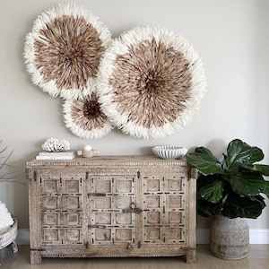 Peut inclure: Trois décorations murales rondes avec un centre marron et des bords en plumes blanches sont suspendues au-dessus d'un meuble en bois. Le meuble présente des détails sculptés et est surmonté d'objets décoratifs. Une grande plante en pot est à droite.