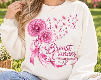 May include: A cream-colored crewneck sweatshirt featuring a graphic print of two pink dandelion heads and a pink awareness ribbon. The design includes floating pink dandelion seeds and the text Breast Cancer Awareness in pink script lettering.