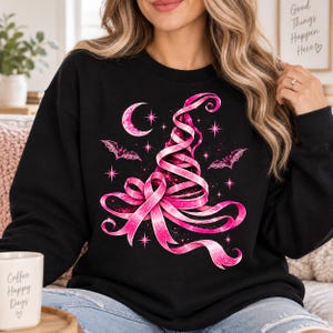 May include: A black long-sleeve sweatshirt featuring a graphic print of a witch hat constructed from a pink ribbon. The design includes a crescent moon, two small bats, and several four-pointed stars, all rendered in shades of bright pink.
