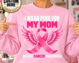 May include: A light pink crewneck sweatshirt featuring the white and pink text I WEAR PINK FOR MY MOM and BREAST CANCER AWARENESS. The design includes a large pink awareness ribbon with detailed pink wings extending from the sides of the ribbon.