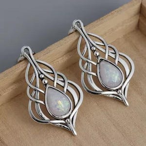 Puede incluir: Un par de pendientes de plata con una piedra blanca iridiscente en el centro de cada pendiente. Los pendientes tienen un diseño delicado e intrincado.
