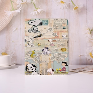 May include: A rectangular notebook featuring Snoopy and other Peanuts characters. The design includes comic strip panels, stars, and the text "COSMIC CONNECTION!". The color palette is a mix of beige, blue, and white.