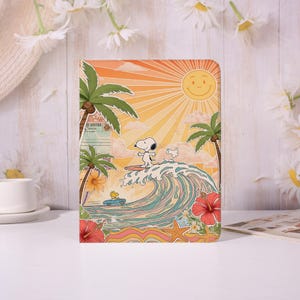 May include: A colorful notebook featuring Snoopy surfing a wave under a smiling sun. Palm trees, hibiscus flowers, and a vintage car add to the beach theme. The background has an orange and yellow gradient.