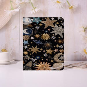 May include: Black tablet case with a celestial design featuring gold, silver, and blue stars, moons, and suns. The case has a textured, beaded appearance, creating a detailed and eye-catching pattern. The design is set against a black background.
