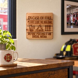 Könnte beinhalten: Ein quadratisches Wandschild aus Holz mit dem eingravierten Text IN CASE OF FIRE PLEASE LEAVE THE BUILDING, gefolgt von Symbolen eines Smartphones, eines Feuers und drei Ausrufezeichen sowie dem Text BEFORE POSTING IT ON SOCIAL MEDIA.