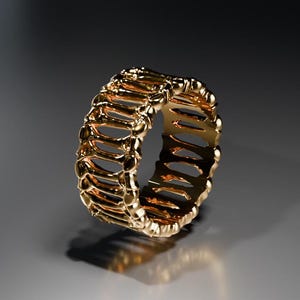 Può includere: Un anello color oro con un design traforato unico. L'anello presenta una serie di barre verticali collegate da bande orizzontali, creando un motivo intricato. L'anello è esposto su una superficie riflettente, evidenziando la sua lucentezza metallica.