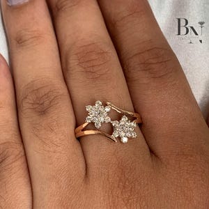 May include: A rose gold ring featuring two flower-shaped clusters of small, sparkling diamonds. The ring's band splits to cradle each floral design, creating a delicate and elegant look. The ring is shown on a hand with a neutral nail polish.