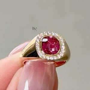 May include: A gold ring with a large, square-cut, deep red gemstone encircled by a halo of small, clear diamonds. The ring's band is smooth and polished. The ring is held up by a person's fingers.