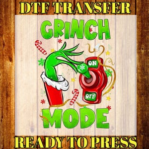 May include: A DTF transfer design with the words "GRINCH MODE" in green, featuring a Grinch hand, a red switch, and candy canes. The text "READY TO PRESS" is also included. The design is on a light wood grain background.