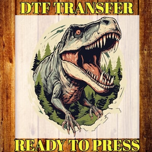 May include: A DTF transfer featuring a detailed illustration of a Tyrannosaurus Rex with an open mouth, revealing sharp teeth. The dinosaur is gray and red, set against a green forest backdrop. The text "DTF TRANSFER" and "READY TO PRESS" are in yellow.