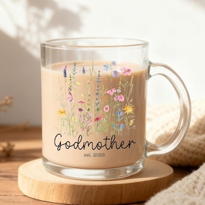 Puede incluir: Una taza de vidrio transparente con asa, llena de un líquido marrón claro. La taza está decorada con un diseño floral en varios colores y las palabras "Godmother est. 2025". La taza está sobre un posavasos de madera.