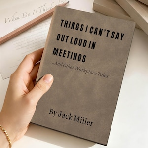 Puede incluir: Un libro gris con el título "THINGS I CAN'T SAY OUT LOUD IN MEETINGS...And Other Workplace Tales" de Jack Miller. Un lápiz y una hoja de papel con escritura cursiva son visibles en el fondo. El libro está sostenido por una persona.