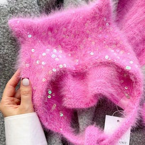 Puede incluir: Un gorro de punto rosa brillante y esponjoso con puntas en forma de oreja y una cinta. El sombrero está adornado con lentejuelas dispersas. El sombrero lo sostiene una persona que lleva una camisa blanca.