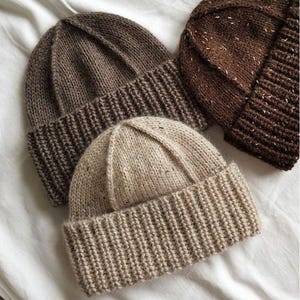 May include: Three knitted beanies in shades of brown and beige are displayed on a white surface. The beanies have a ribbed brim and a textured knit pattern. The colors range from light beige to a dark brown, with one beanie having flecks of white.