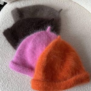 Può includere: Una collezione di quattro morbidi cappelli in maglia pelosa nei toni dell'arancione, rosa, marrone e grigio. Ogni cappello ha un bordo ripiegato e un piccolo dettaglio a punta sulla sommità. I cappelli sono disposti su una superficie bianca sporca.