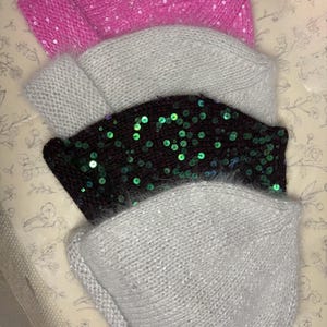 May include: A stack of four knitted beanies in shades of pink, gray, black, and silver. The pink beanie is covered in sequins, while the black beanie has green sequins. The beanies are made of a soft, fuzzy material.