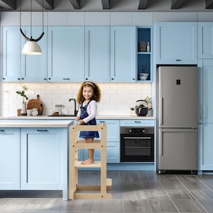 May include: A light wood step stool in a kitchen with pale blue cabinets. A young child stands on the stool, smiling. The kitchen has a stainless steel fridge and a black oven. The room is well-lit with natural light.
