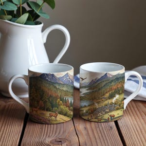 May include: Two white ceramic mugs with a scenic landscape design. The mugs feature a mountain range, lake, and forest scene with deer and an eagle. The mugs are on a wooden surface with a white jug and greenery in the background.