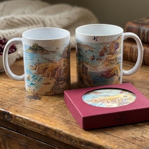 May include: Two white ceramic mugs featuring a colorful watercolor illustration of a coastal landscape with a castle on an island, accompanied by a square maroon gift box containing a matching circular coaster with the same coastal artwork.