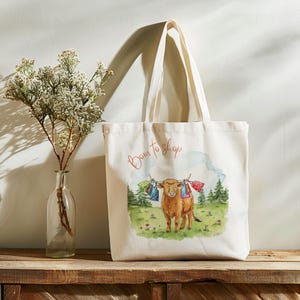 Op de afbeelding: Crèmekleurige draagtas met de tekst "Born to shop" in rood script. De tas heeft een aquarel illustratie van een bruine koe met boodschappentassen die aan haar hoorns hangen, in een groen veld met bomen.