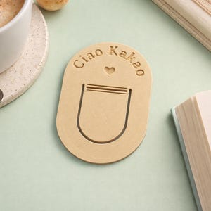 Könnte beinhalten: Ein hellbrauner Lesezeichen mit dem Text "Ciao Kakao" und einem Herzsymbol. Das Lesezeichen hat ein Taschen-Design. Eine Tasse Kaffee, eine Untertasse und ein Löffel sind im Hintergrund, zusammen mit einem Buch.