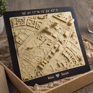 May include: A 3D map of a city, in a beige color, mounted on a black frame. The map is inside a cardboard box with a tag. The coordinates 52 31' 17" N 13° 24' 43" O are at the top. The names "Max" and "Sarah" are at the bottom with a heart symbol.