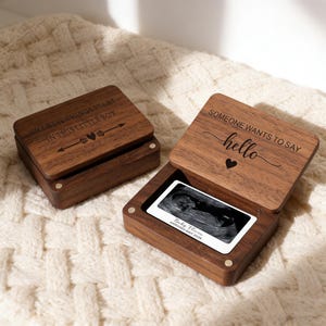 Op de afbeelding: Twee houten bewaardoosjes, één gesloten en één open, met gegraveerde tekst. De open doos toont een echografie. De gesloten doos leest "THE BEST THINGS START IN THIS LITTLE BOX." De open doos leest "SOMEONE WANTS TO SAY hello."