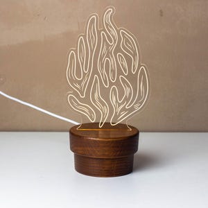 May include: A decorative night light featuring a clear acrylic design of abstract shapes, illuminated by a warm glow. The light sits on a round, dark brown wooden base, with a white power cord extending from the back. The lamp is designed for home decor.