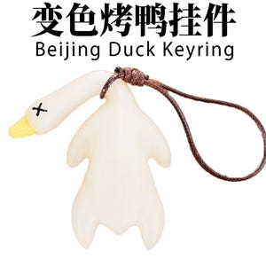 May include: A white Peking duck keyring with a brown cord. The duck has a yellow beak and a black cross on its head. The text "Beijing Duck Keyring" is printed below the duck.