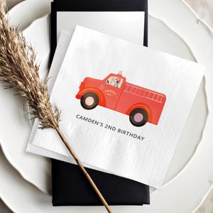 May include: White square paper napkin with a red fire truck illustration and the text "CAMDEN'S 2ND BIRTHDAY". The napkin is placed on a white plate with a black card and a dried plant stem.