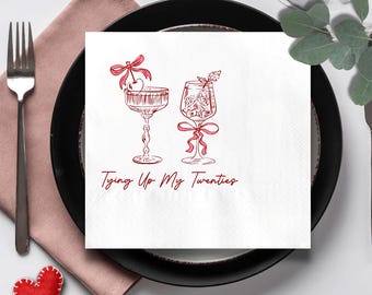 May include: A white cocktail napkin with a red illustration of two cocktail glasses, a cherry, and bows. The text "Tying Up My Twenties" is written in red script below the glasses. The napkin is on a black plate with a pink napkin and a fork.