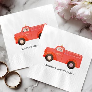 May include: Two white paper napkins with a red fire truck design. The fire trucks have black wheels and a ladder on the back. The text "CAMDEN'S 2ND BIRTHDAY" is printed below the fire truck.