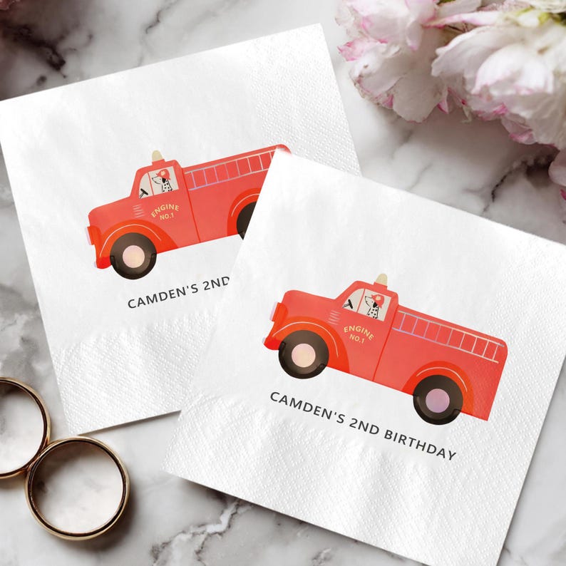 May include: Two white paper napkins with a red fire truck design. The fire trucks have black wheels and a ladder on the back. The text "CAMDEN'S 2ND BIRTHDAY" is printed below the fire truck.