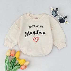 May include: Cream-colored baby romper with the text "PASS ME TO grandma" in black script and a red heart. The long-sleeved romper is paired with sunglasses and a bouquet of tulips.