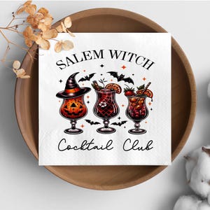 May include: White square napkin with the text "Salem Witch Cocktail Club" and illustrations of three cocktails. The first cocktail is in a glass shaped like a jack-o'-lantern. The other two cocktails have fruit garnishes.