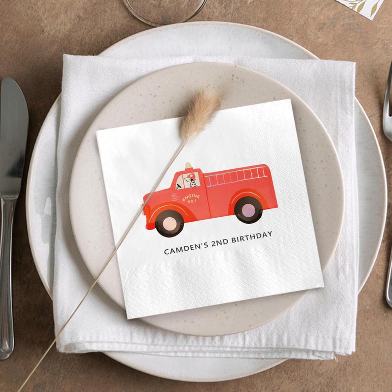 May include: White square napkin with a red fire truck illustration. The fire truck has black wheels and the text "ENGINE NO. 1". The napkin also has the text "CAMDEN'S 2ND BIRTHDAY". The napkin is placed on a white plate with a white napkin and silverware.