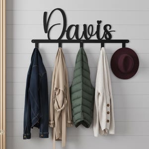 Pode incluir: Um cabide de parede de metal preto com o nome "Davis" em escrita cursiva. O cabideiro suporta uma jaqueta jeans, um trench coat bege, um colete acolchoado verde, um cardigan creme e um chapéu bordô. O cabideiro é montado em uma parede cinza com ripas horizontais.