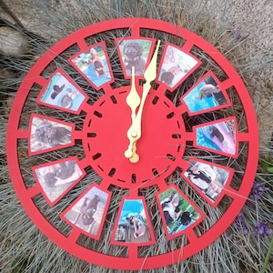 Könnte beinhalten: Eine runde, rote Uhr mit dekorativem Außenring und goldfarbenen Zeigern. Das Zifferblatt zeigt kleine Fotorahmen mit Bildern von Hunden. Die Uhr liegt auf einem Grasbett.