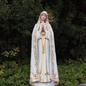 Op de afbeelding: Een wit en goud beeld van een religieuze figuur, met de handen gevouwen in gebed. De figuur draagt een witte jurk en een lichtblauwe mantel met gouden randen. Het beeld staat tegen een achtergrond van groen gebladerte.