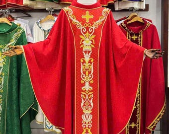 May include: A red liturgical vestment with gold embroidery, displayed on a mannequin. The vestment features a cross and ornate designs. Other colorful vestments, including green, red, and purple, are visible in the background, suggesting a religious theme.